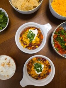 2 bowls of vegetarian chili topped with Greek yogurt, shredded cheese, sour cream and cilantro. With extra side toppings on the side.