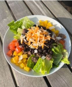 taco bowl on a picnic table with lettuce, peppers, tomatoes, black beans and shredded cheese on top