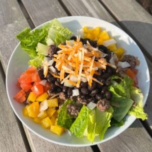 taco bowl on a picnic table with lettuce, peppers, tomatoes, black beans and shredded cheese on top